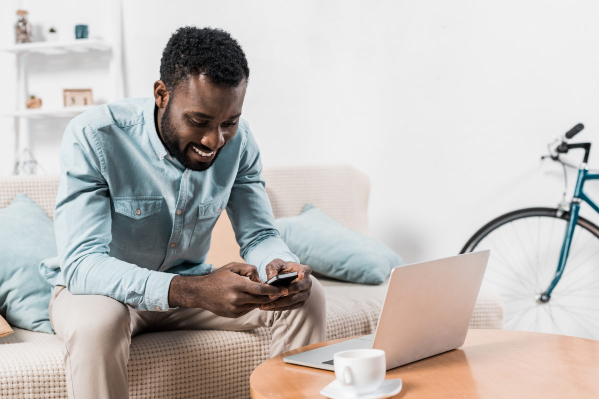 african american man sitting on couch and typing on smartphone