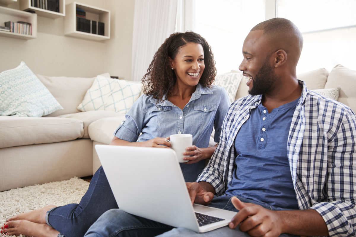 Couple sitting on the ground in front of couch talking, while holding a computo